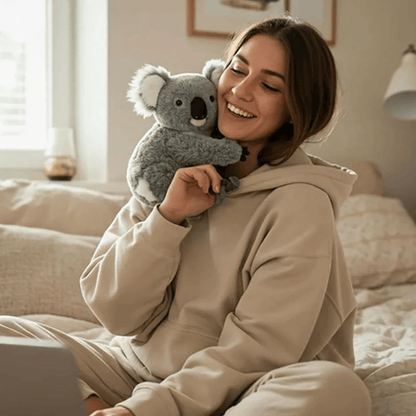 Peluche koala réaliste autour du cou d'une femme assise en tailleur sur un lit 